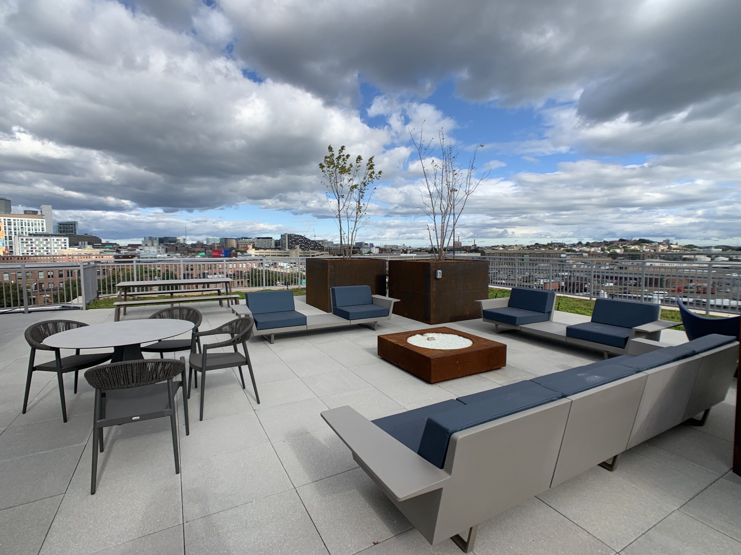 Rooftop deck with modern lounge seating in navy and gray, a fire pit table, dining table with chairs, and planters, overlooking a city skyline under a partly cloudy sky.