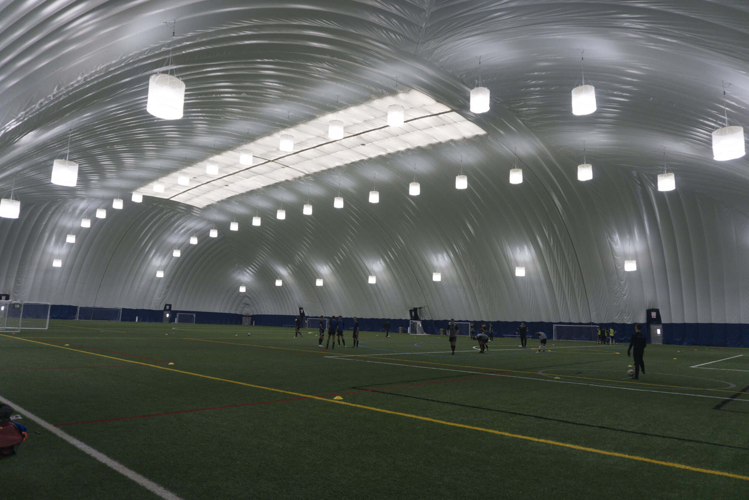 Interior of an air-supported dome facility with a turf soccer field, players training under rows of hanging lights beneath the arched fabric ceiling.