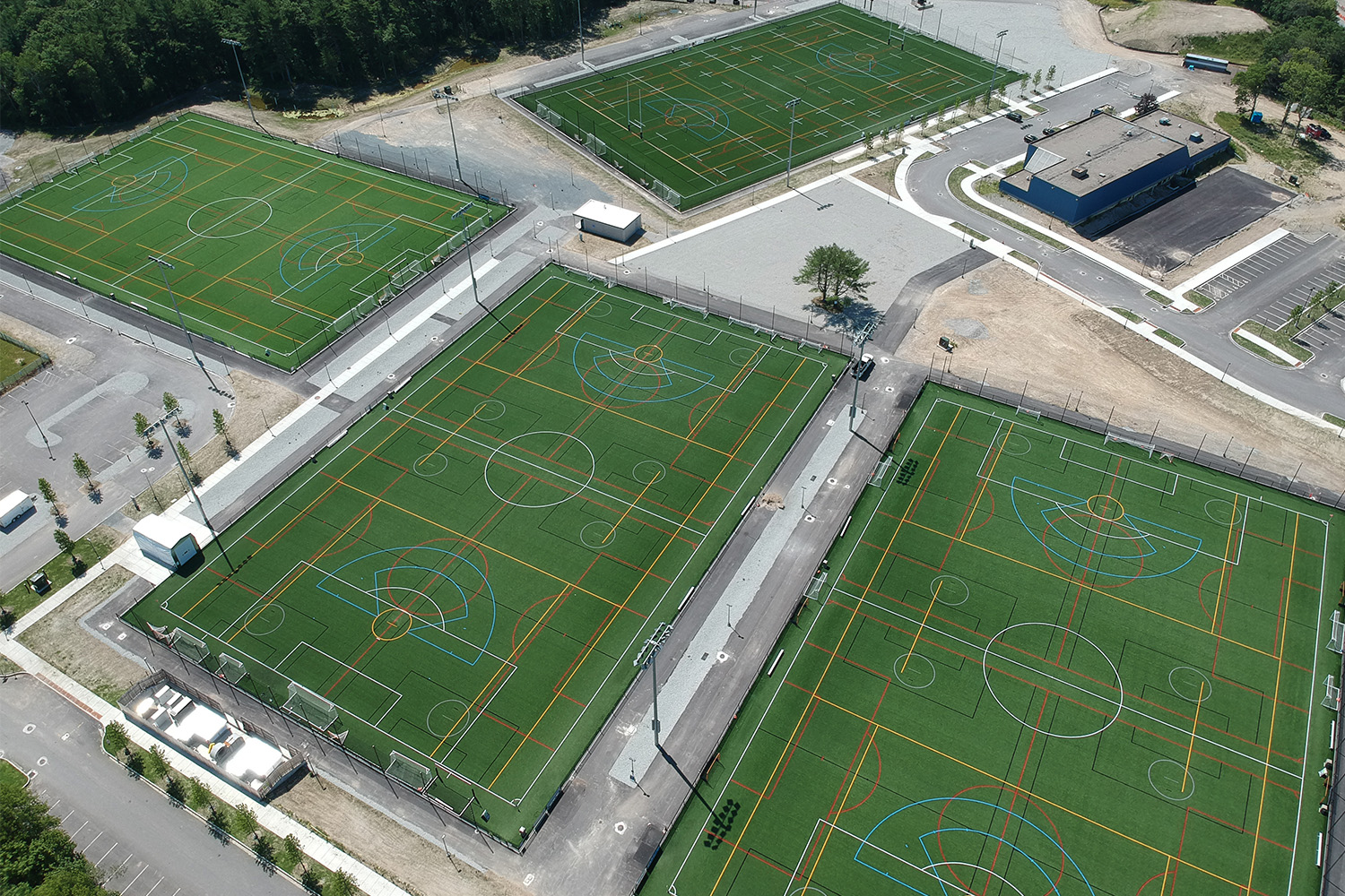 Aerial view of four multi-sport turf fields with overlapping soccer, lacrosse, and field markings, surrounded by parking lots, a maintenance building, and wooded areas.