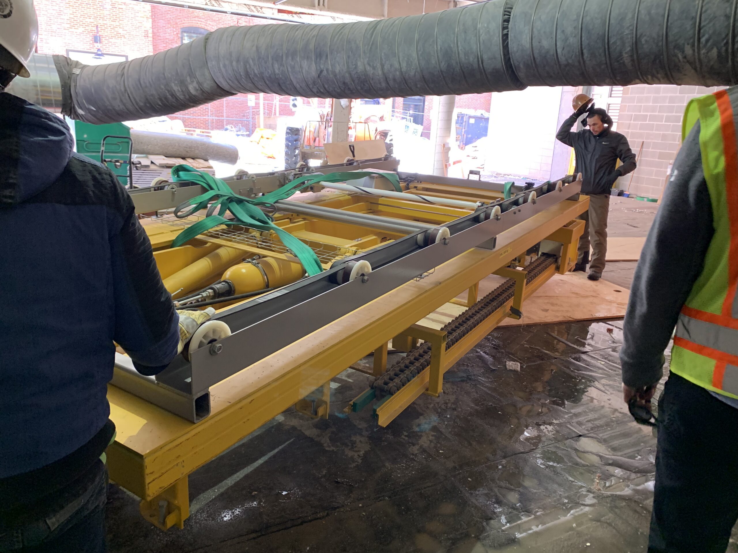 Construction workers in hard hats and safety gear installing a component of an automated parking system, with a yellow rail-mounted carriage secured by lifting straps at an indoor loading area.