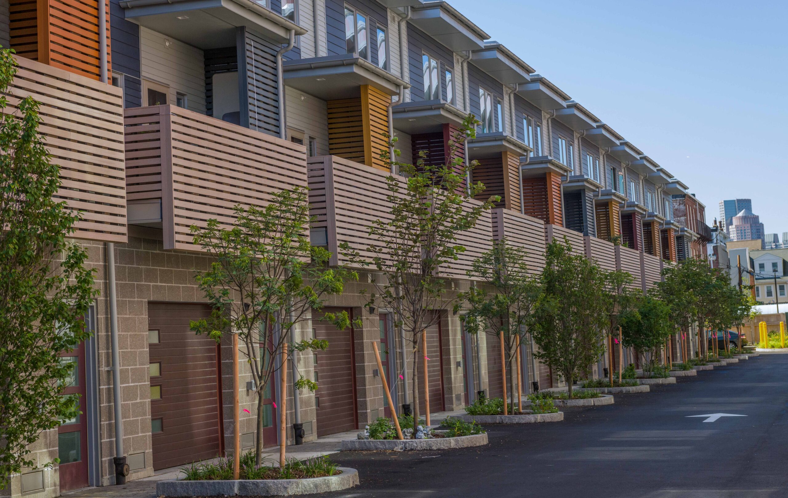 Row of modern townhouses with wood-slat balconies and colorful shutter accents, ground-floor garages, and newly planted trees lining the driveway, with a city skyline visible in the distance.