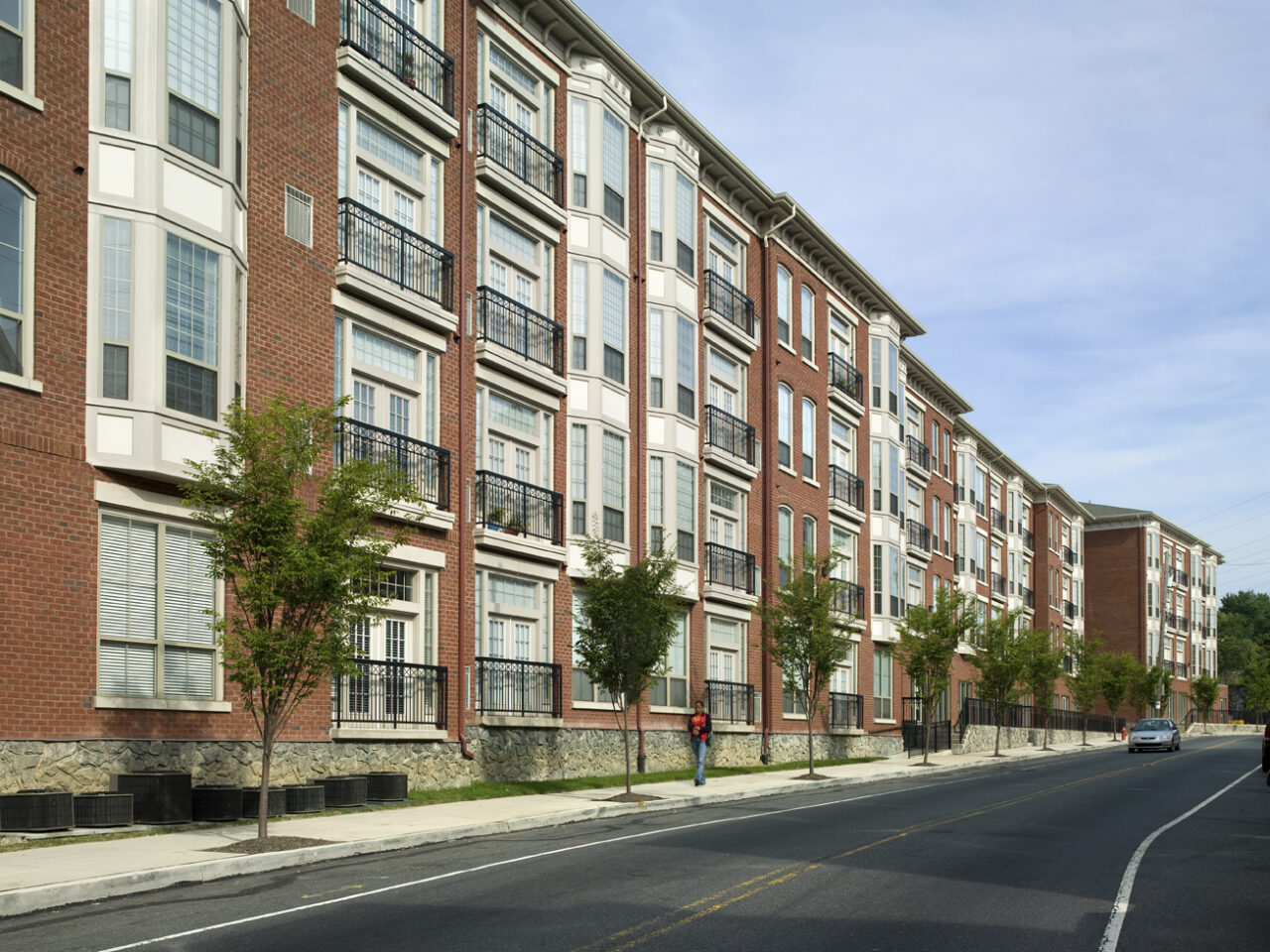 Row of brick apartment buildings with bay windows and iron balconies stretching along a tree-lined street, with a pedestrian and passing car.