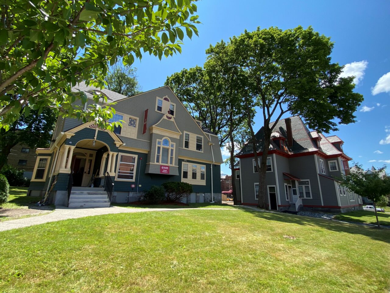 Exterior view of WPI Cedar House B and a neighboring gray Victorian residence hall, showing covered porches, gabled roofs, and a shared front lawn shaded by mature trees.