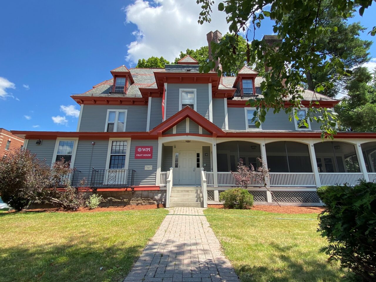 Exterior of WPI Fruit House, a large gray Victorian residence hall with red trim, dormer windows, a wraparound screened porch, and a brick walkway leading to the entrance.