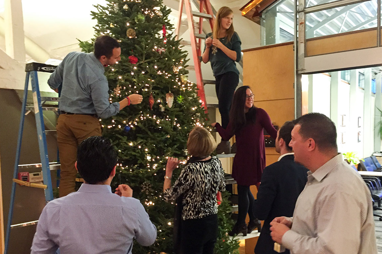 Annual tree trimming at Tocci's headquarters. Staff gather around with ornaments, some standing on ladders to decorate the higher parts of the tree