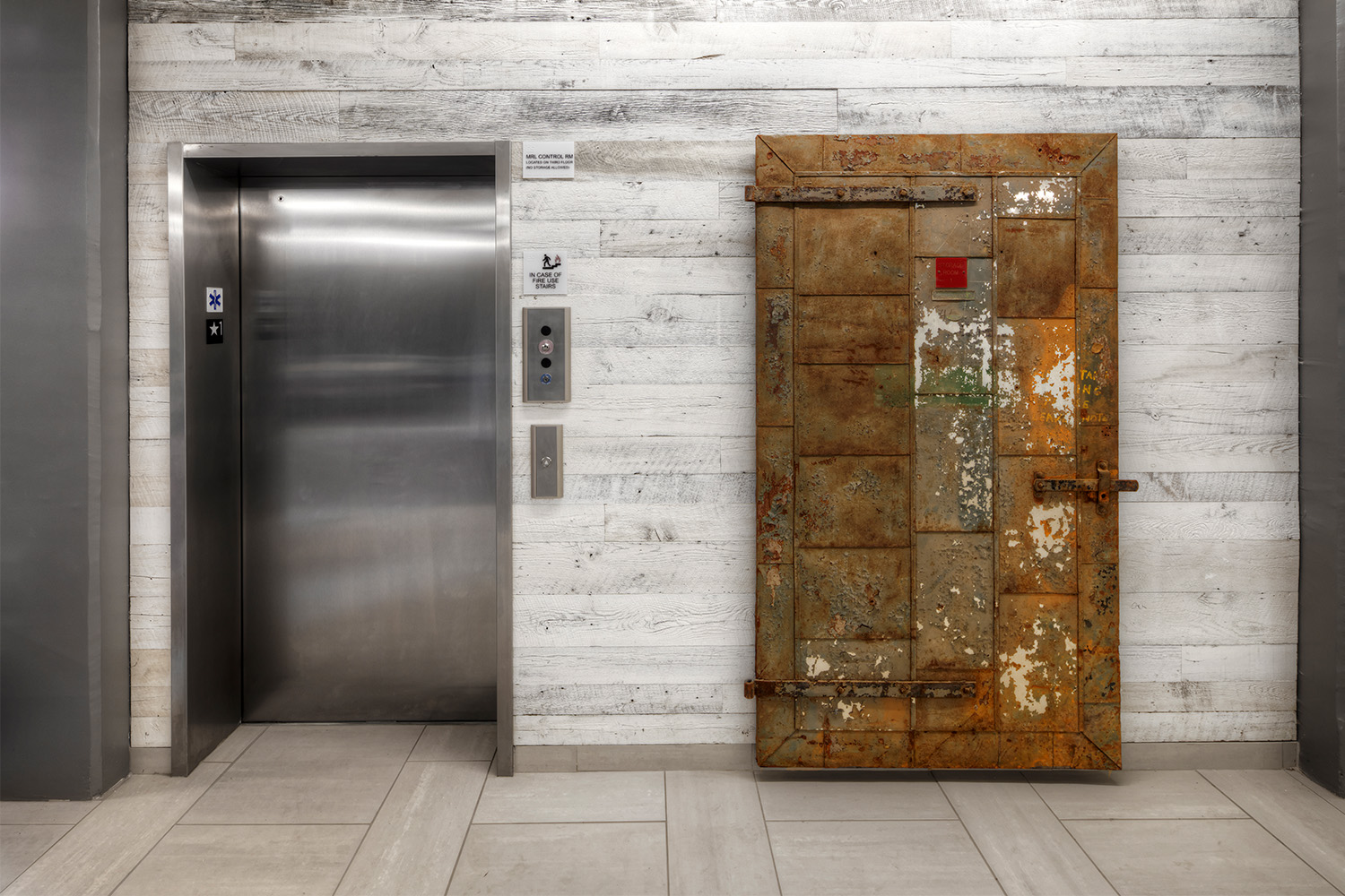 Wall with stylish grey oakwood paneling, an elevator shaft to the left, and a rustic metal door to the right