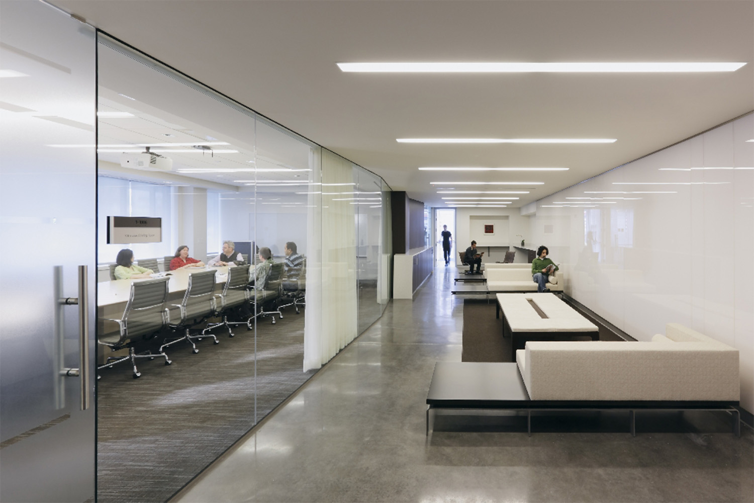 Lounge area with minimalist style white couches and light fixtures, next to occupied conference room 