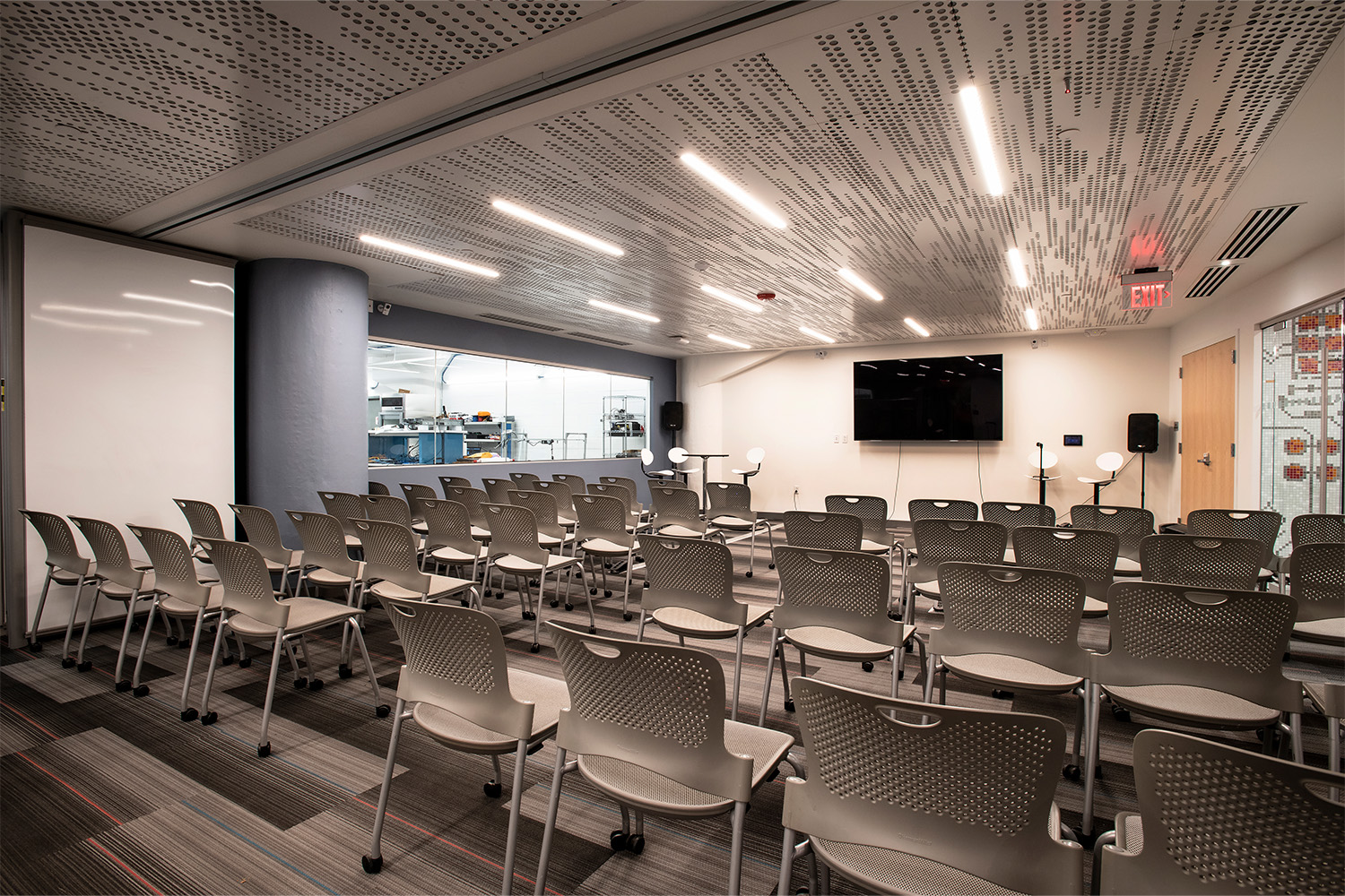 Conference room with rows of grey plastic chairs, TV mounted to wall in front, and long light fixtures on ceiling