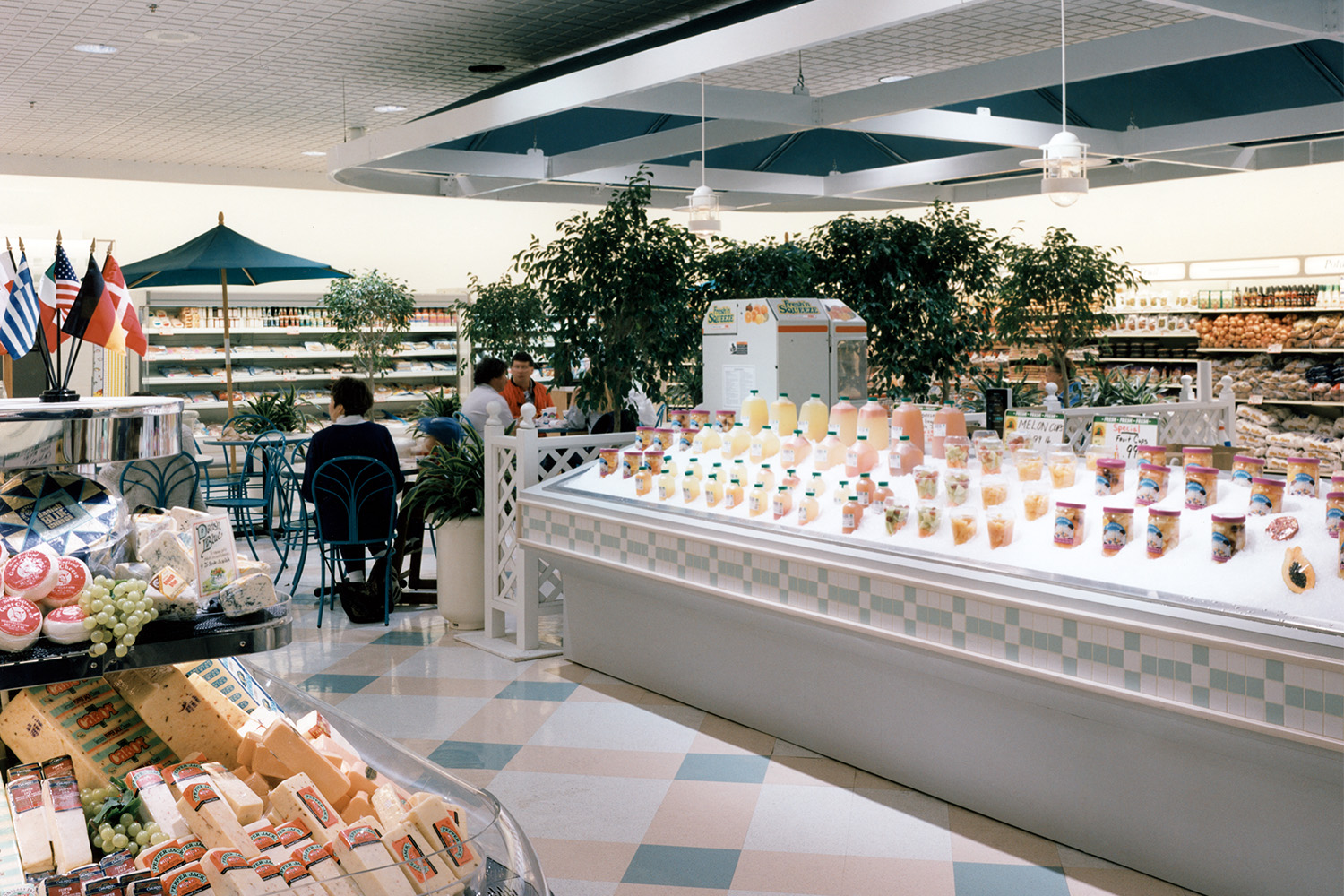 Section of grocery store with juices and fruits on ice to the right, and international cheese section to the left 