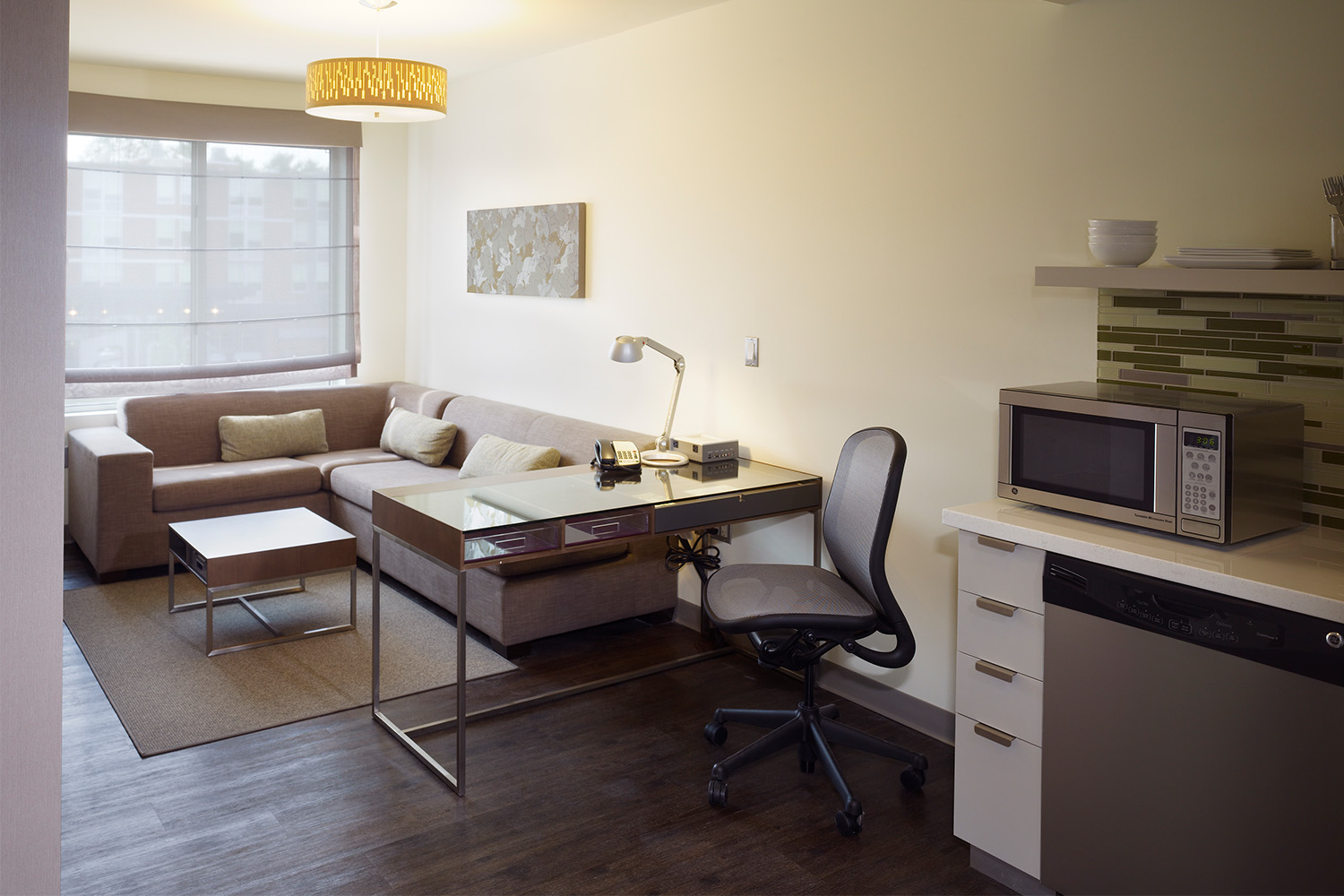 Section of hotel room interior: tan couches and work area, in front of wide window 