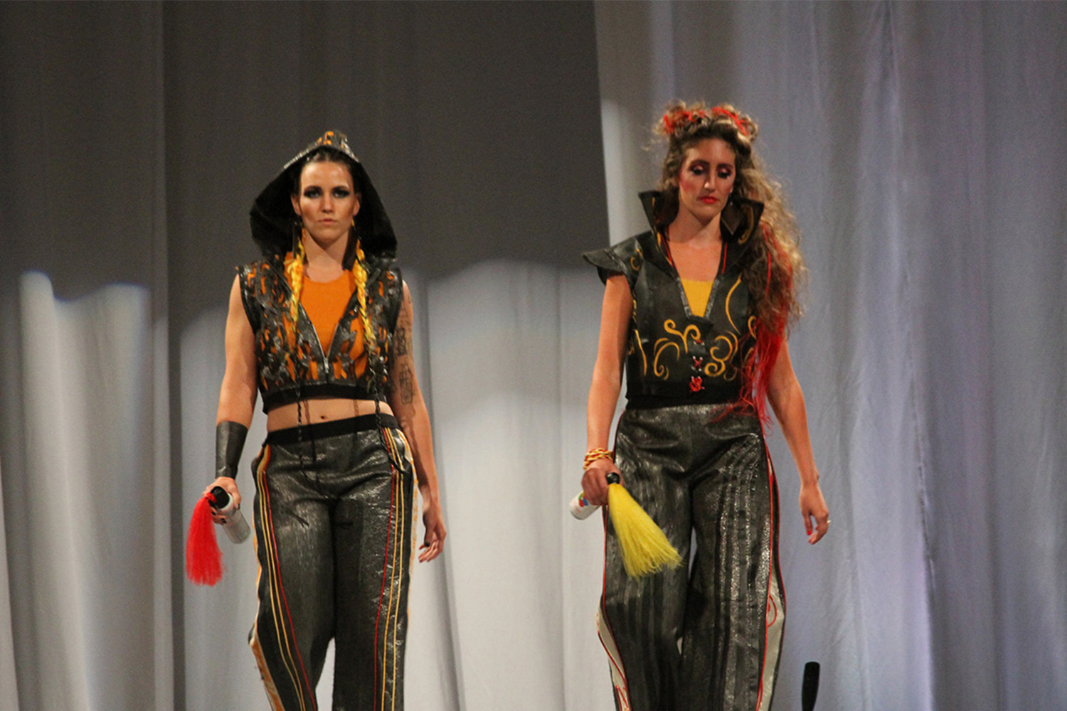 2 female models walking side-by-side down runway 
