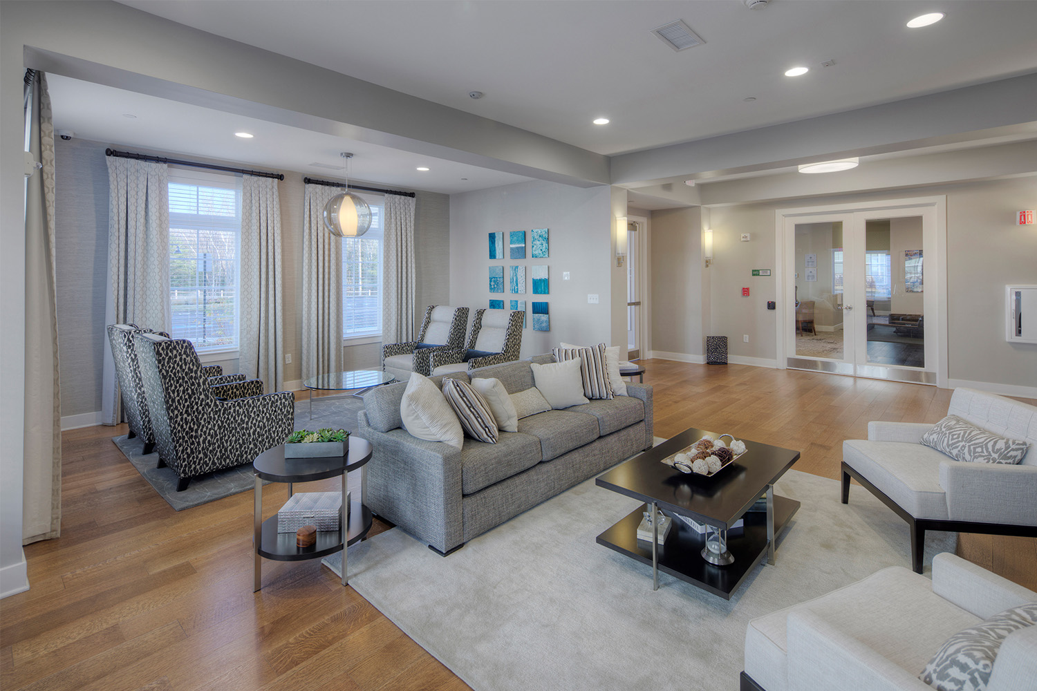 Living room with comfortable grey couches, wooden flooring, and 2 large windows with drapes