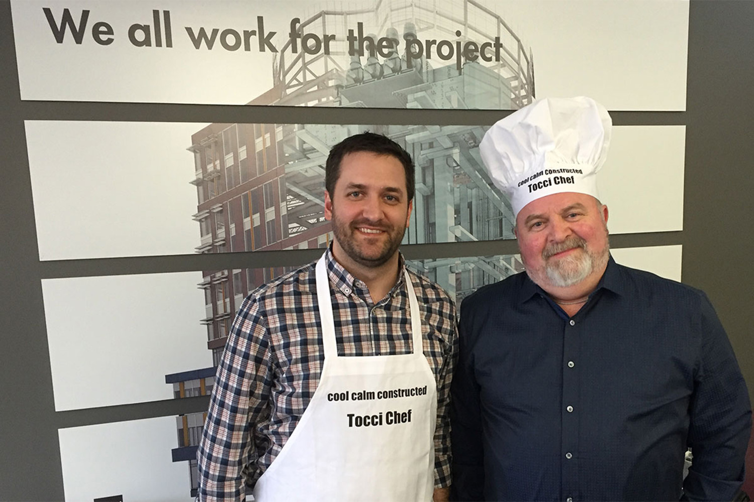 Man wearing apron, next to man wearing chefs hat, both posing