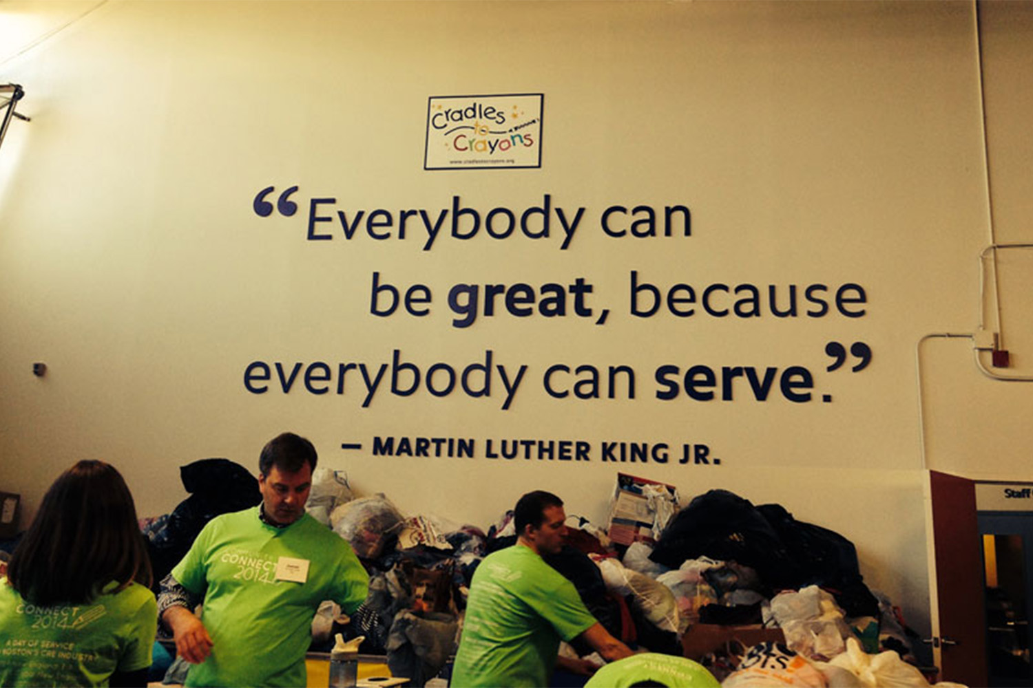 Tocci volunteers sorting through donated clothes, with sigh saying "Everybody can be great, because everybody can serve" on the wall 