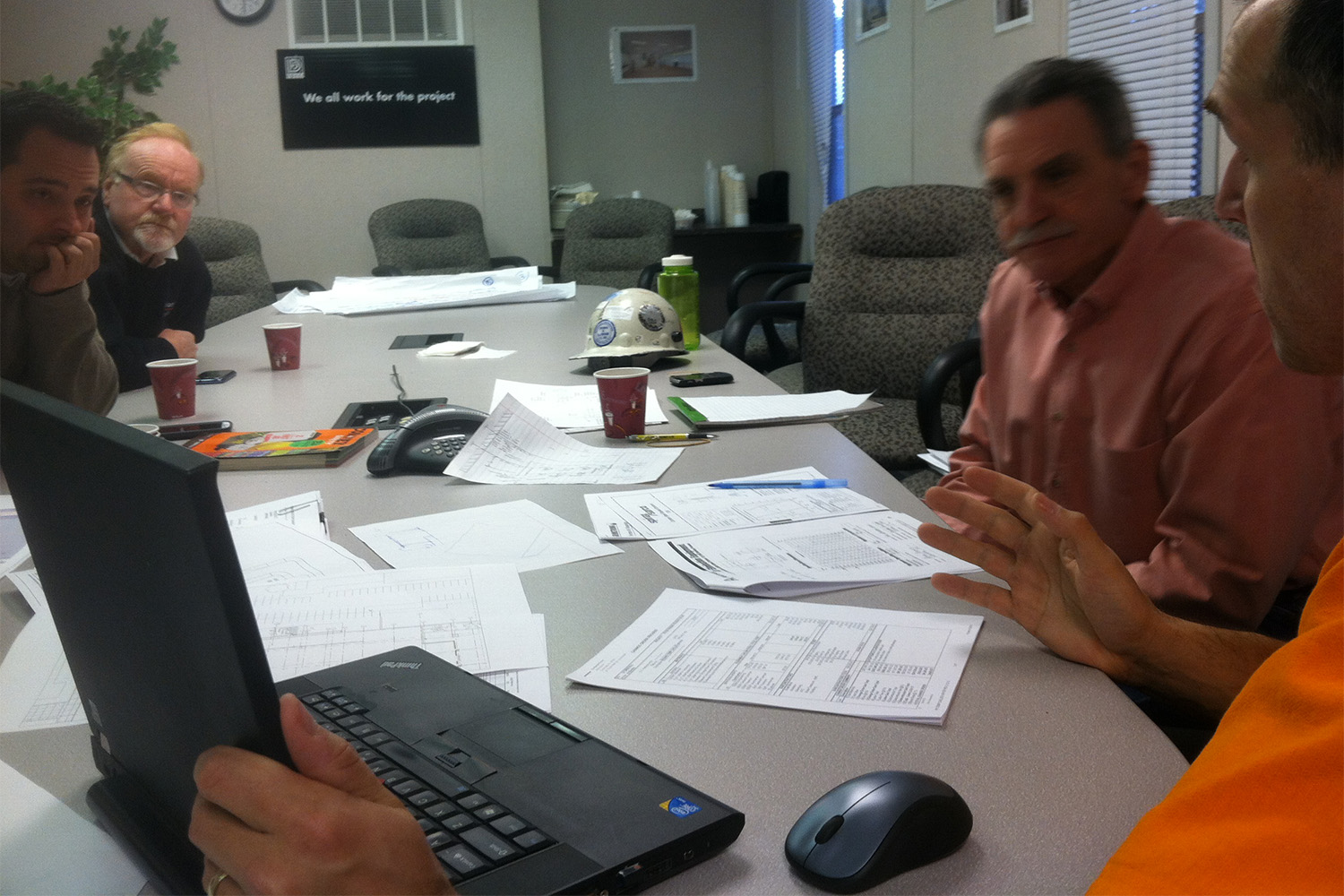 4 men sitting at a table, with the man in an orange shirt showing the others a model on his laptop