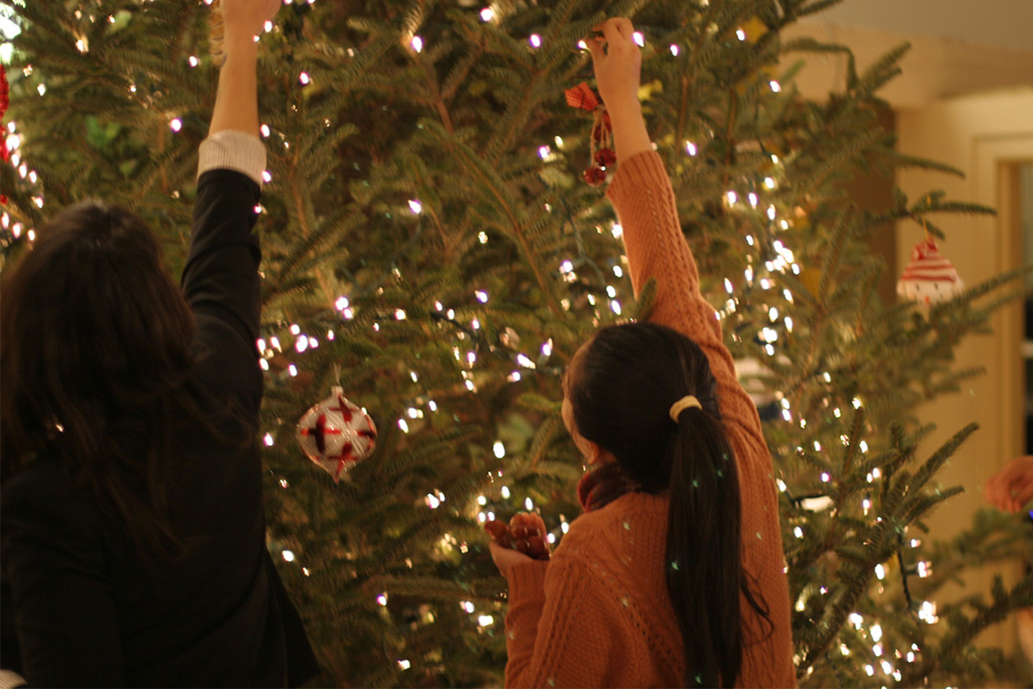 2 people reach to place ornaments on high spot of Christmas tree 