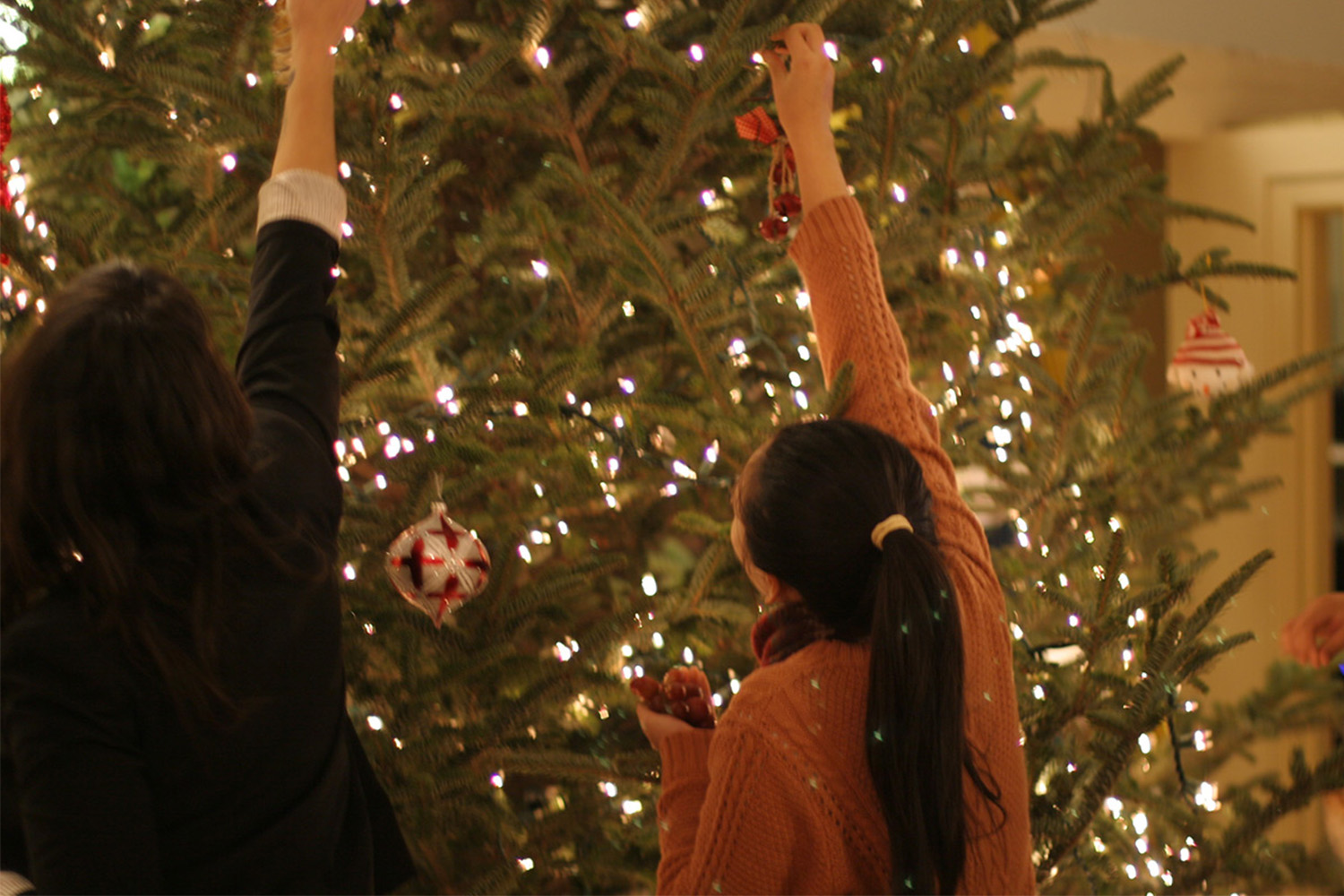 2 people reach to place ornaments on high spot of Christmas tree