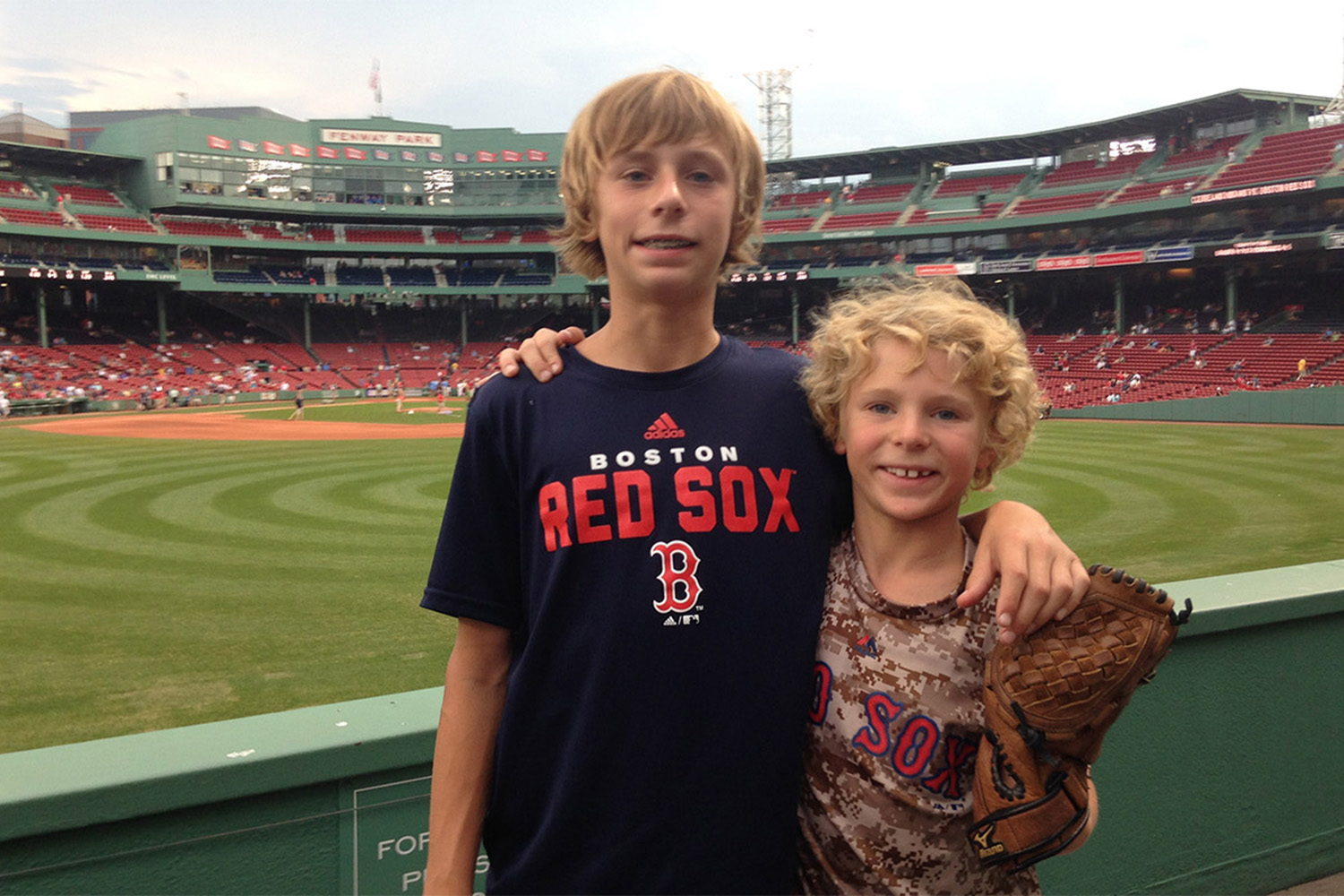 two boys in Red Sox shirts, smiling for photo in front of the Green monster