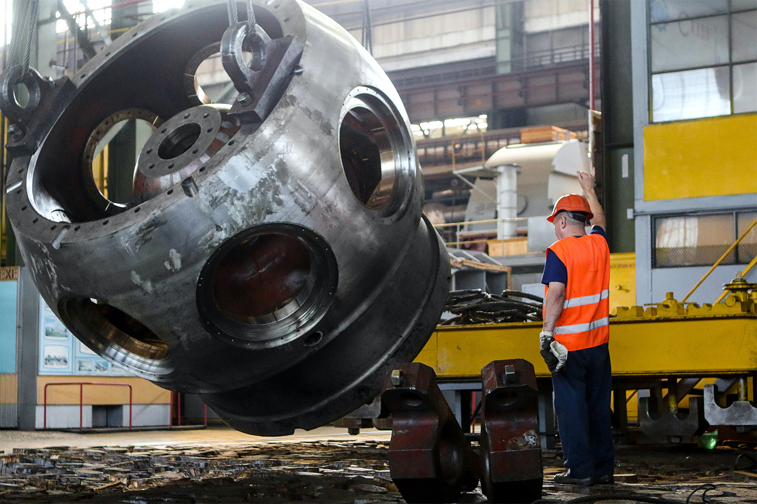 a man in a orange PPE suit, using a machine to lift massive metal object 