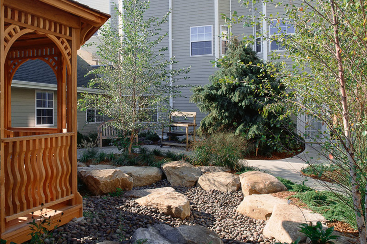 wooden gazebo with rock wall circling it, in front of houses 