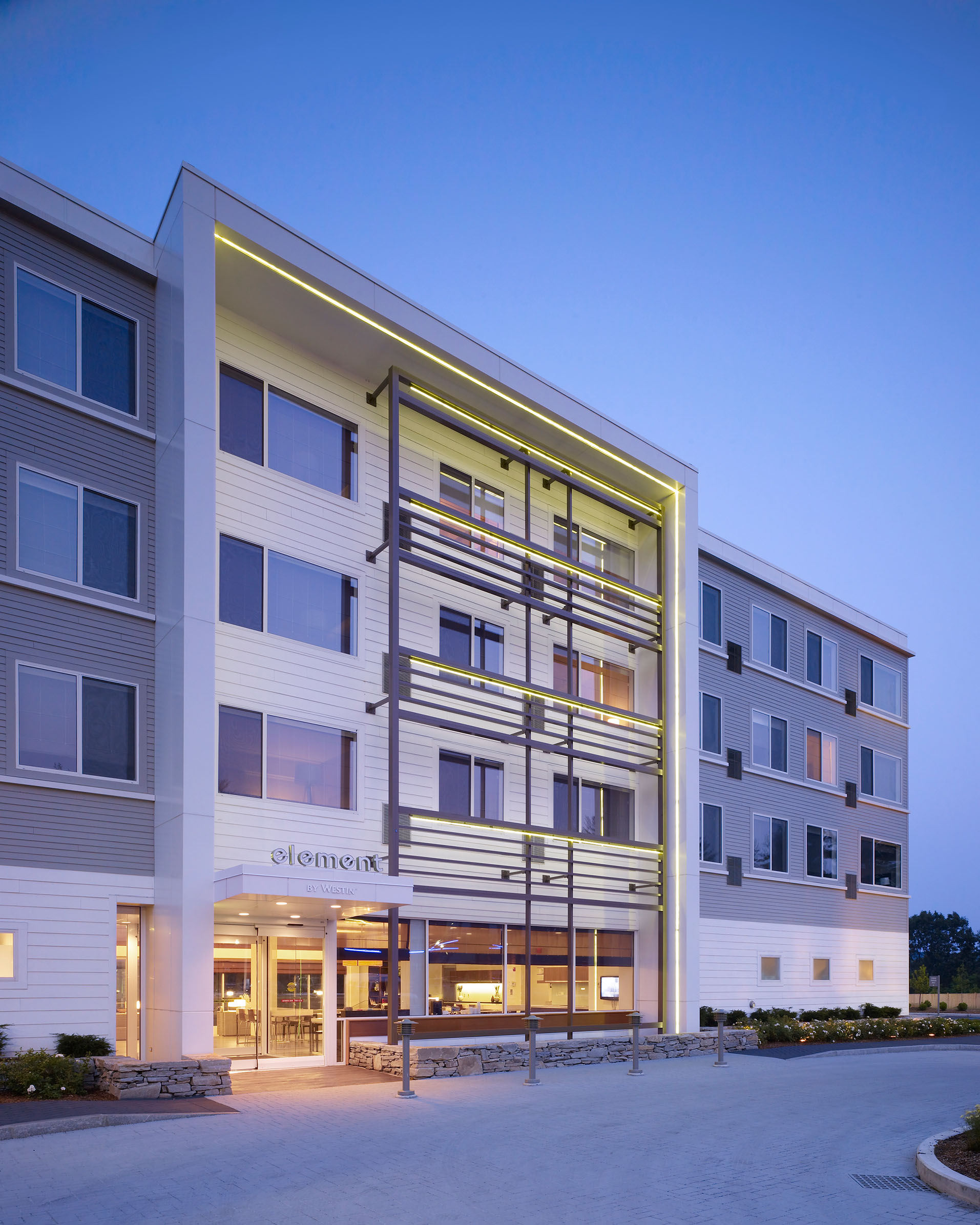 Element by Westin hotel exterior at dusk, illuminated with accent lighting along a modern architectural frame above the entrance, with a stone retaining wall and paved driveway in front.