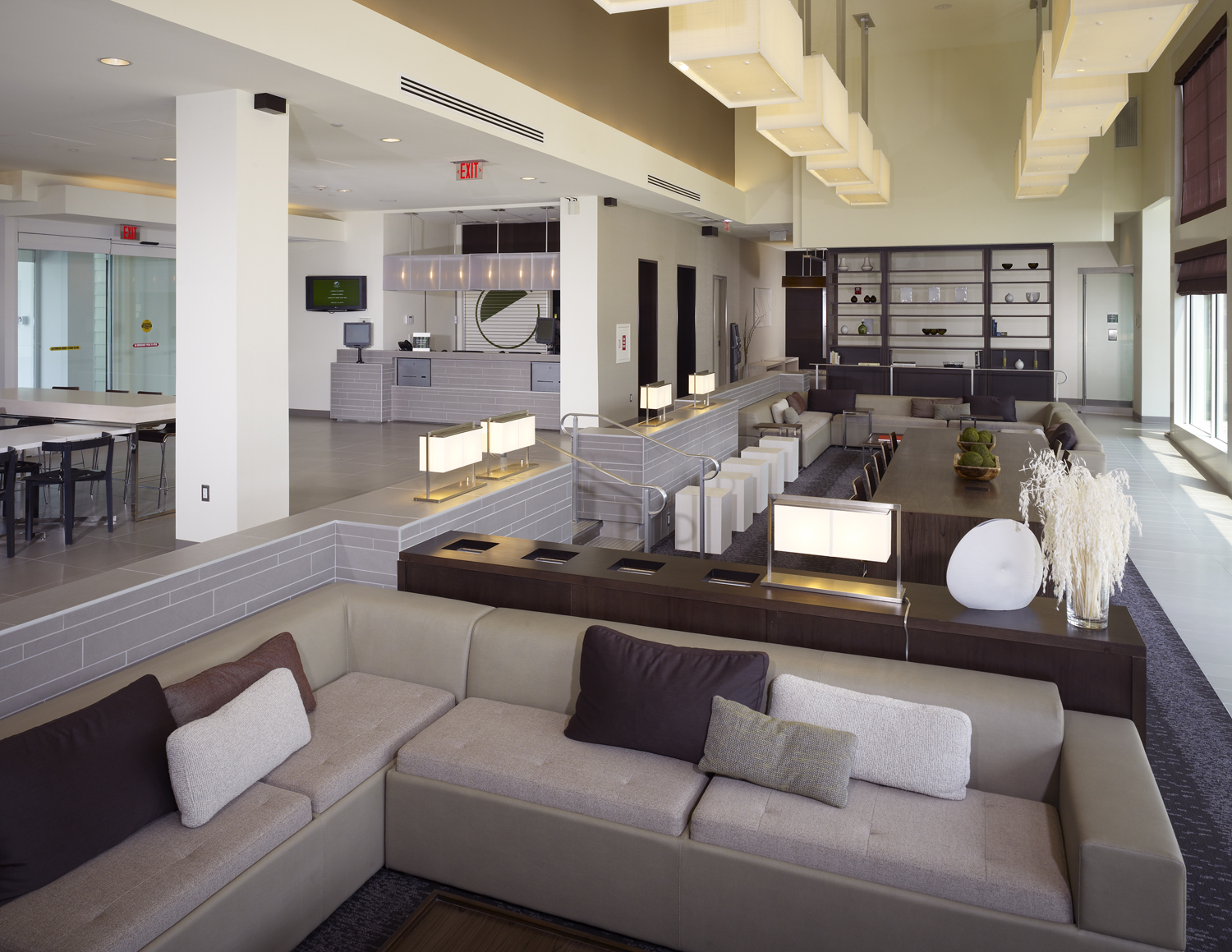 Hotel lobby lounge with a gray sectional sofa, decorative table lamps, a communal dining table, and a front desk with a decorative shelving display in the background.