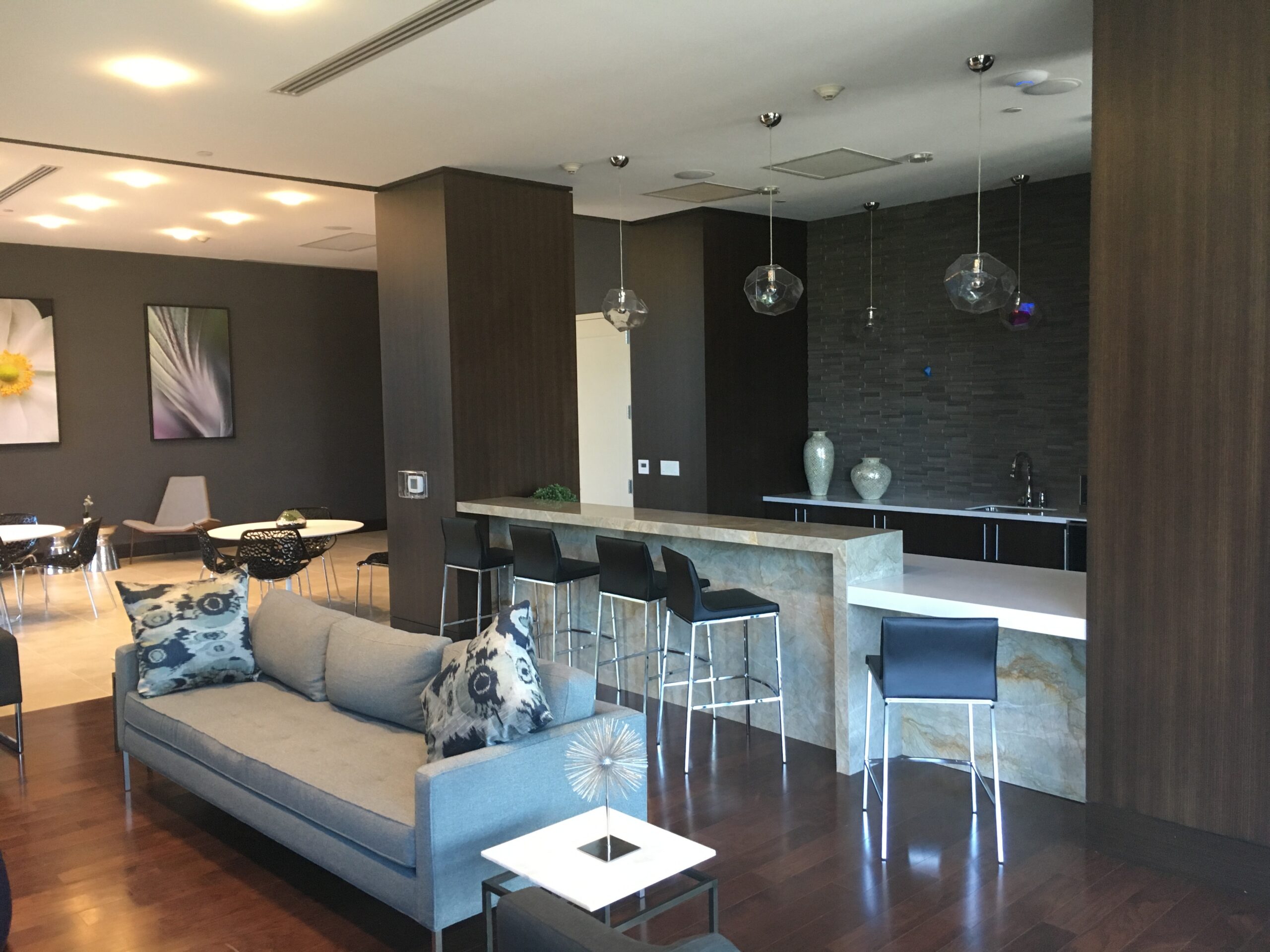 a residential lounge with a gray sectional sofa, a marble bar with black stools, geometric glass pendant lights, and a dark stacked-stone accent wall with a wet bar.