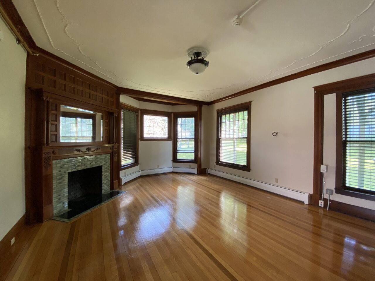 Empty living room with a carved wood fireplace mantel and tiled hearth, a bay window with stained glass accents, and hardwood floors.