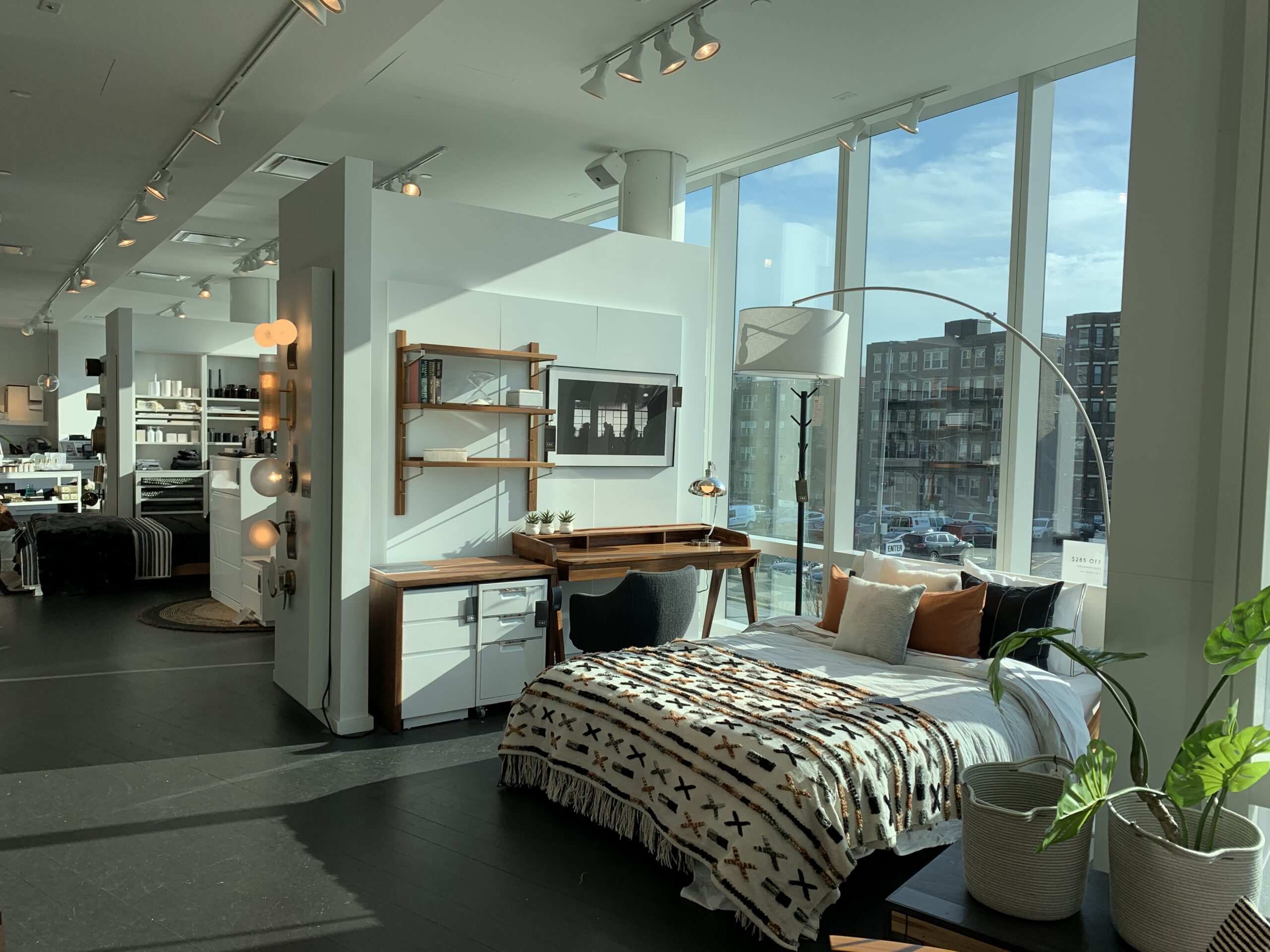 CB2 Interiors bedroom display with a wood desk and shelving unit, a bed styled with patterned bedding, and floor-to-ceiling windows overlooking the street.