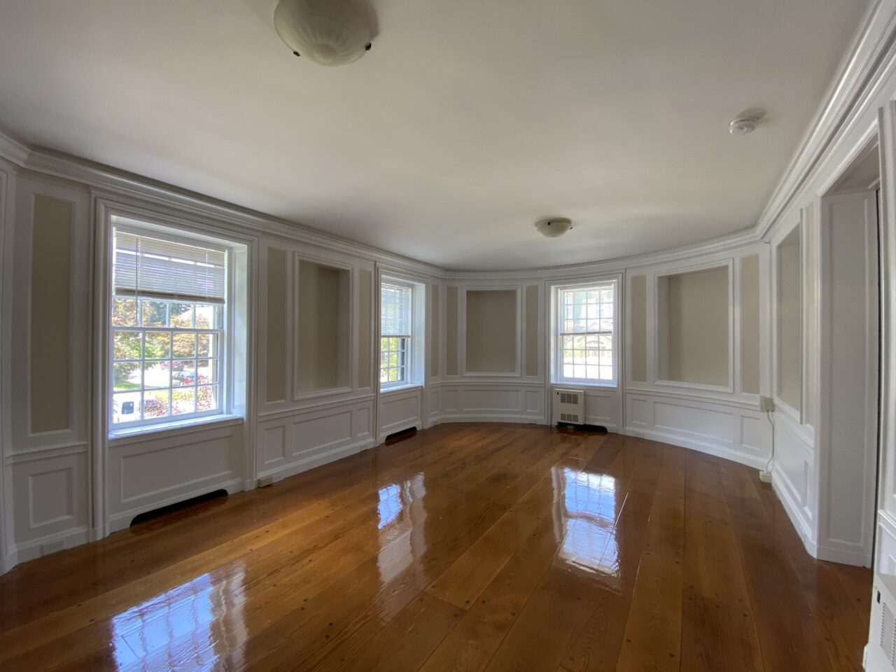 Empty room with a curved bay wall, tall multi-pane windows, decorative wood wainscoting and wall panels, and glossy hardwood flooring.