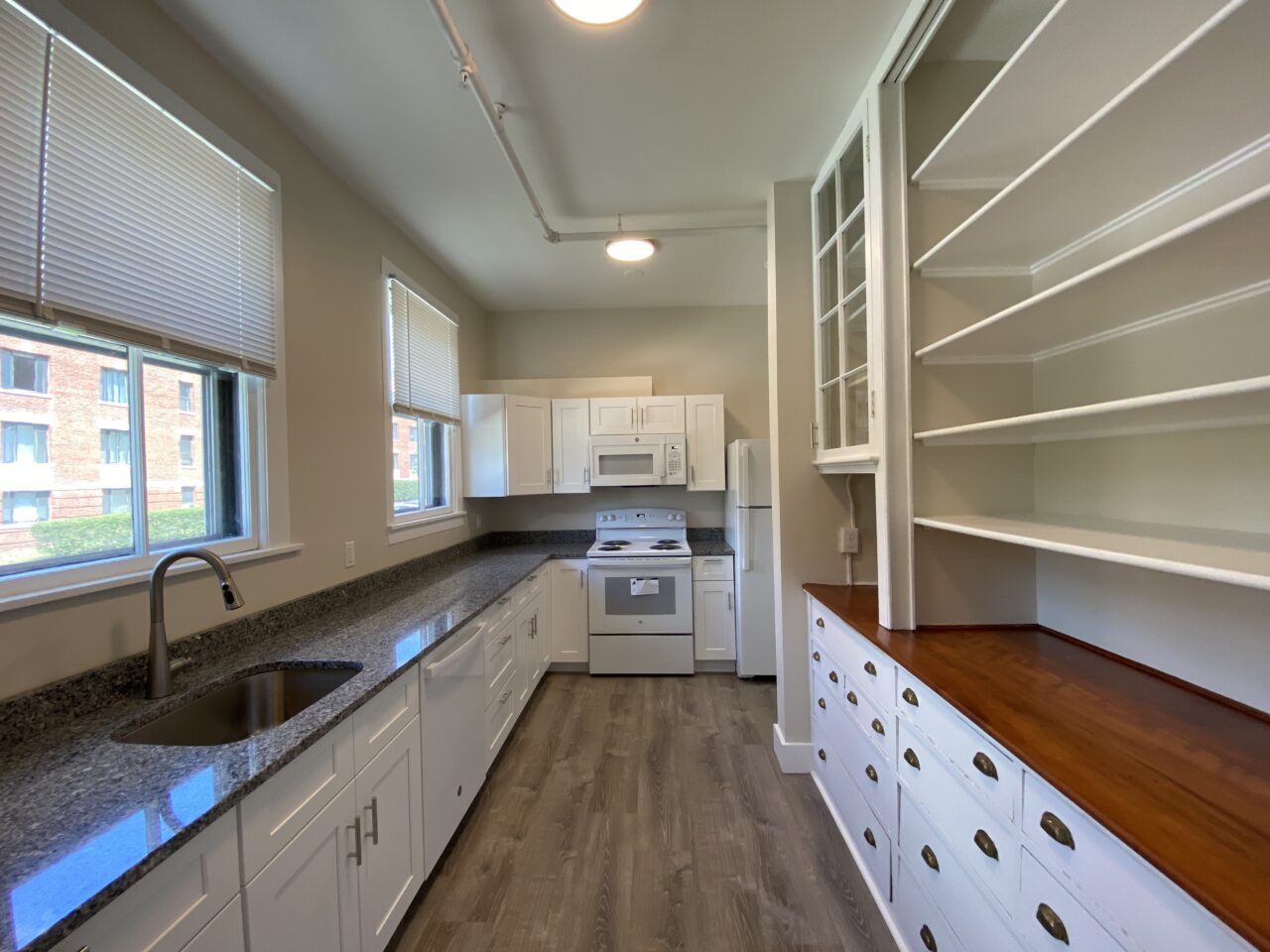 Kitchen with white cabinetry, granite countertops, and stainless sink along one wall, white stove, microwave, and refrigerator opposite, and a built-in wood-topped pantry cabinet with open shelving and vintage drawer pulls.