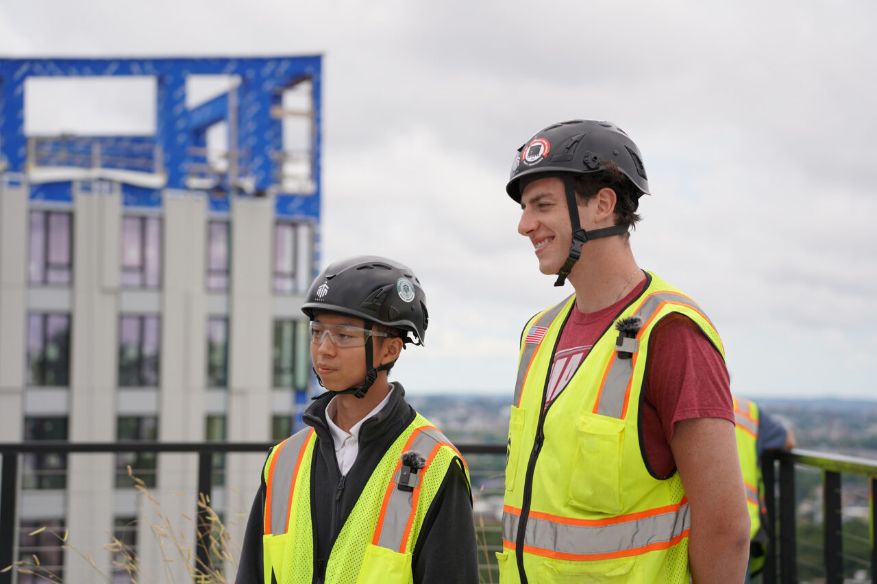 Two workers wearing hard hats and yellow safety vests standing on a rooftop, with an adjacent building under construction in the background.