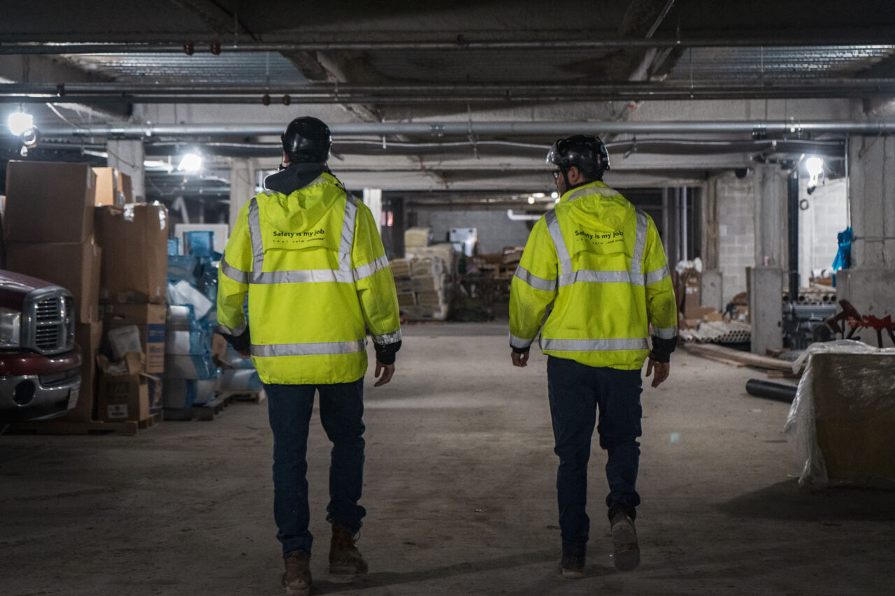 Two workers seen from behind wearing hard hats and yellow safety jackets, walking through a dimly lit underground parking garage lined with stacked construction materials.