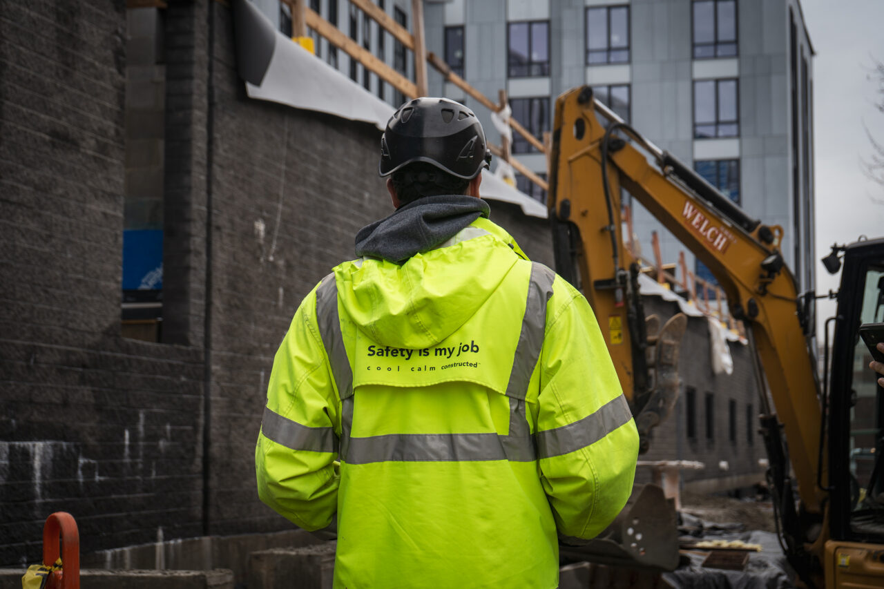 Worker seen from behind wearing a hard hat and yellow jacket standing near an excavator at a construction site with a building rising in the background.