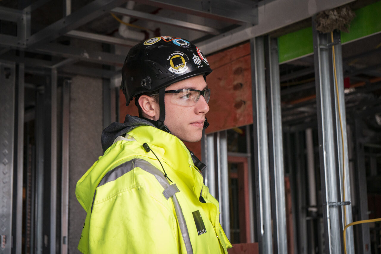 Close-up profile of a worker wearing safety glasses and a sticker-covered hard hat standing inside an unfinished building with exposed metal framing.