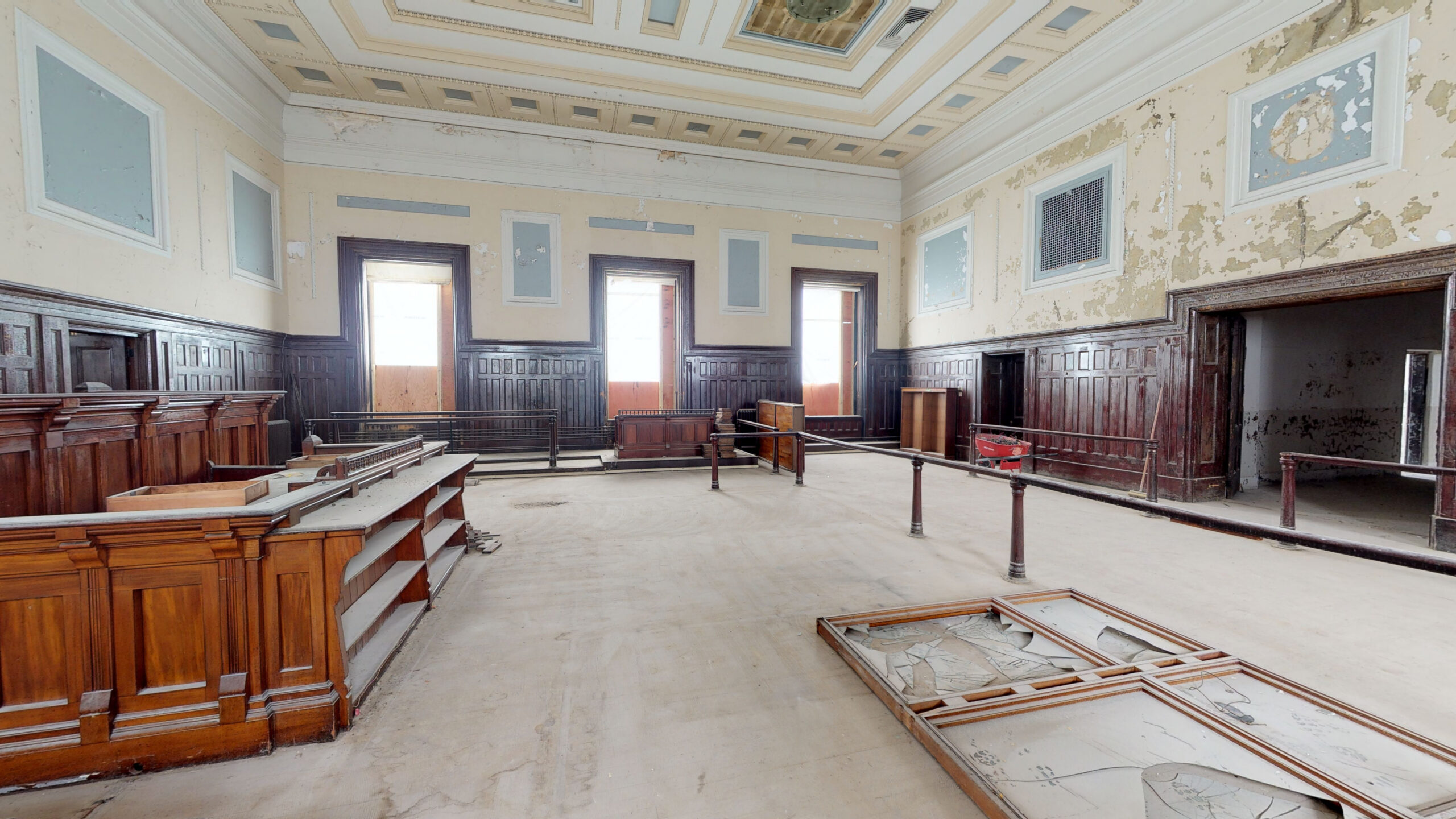 A former courtroom before renovation, showing wood paneling amid peeling paint and construction debris.