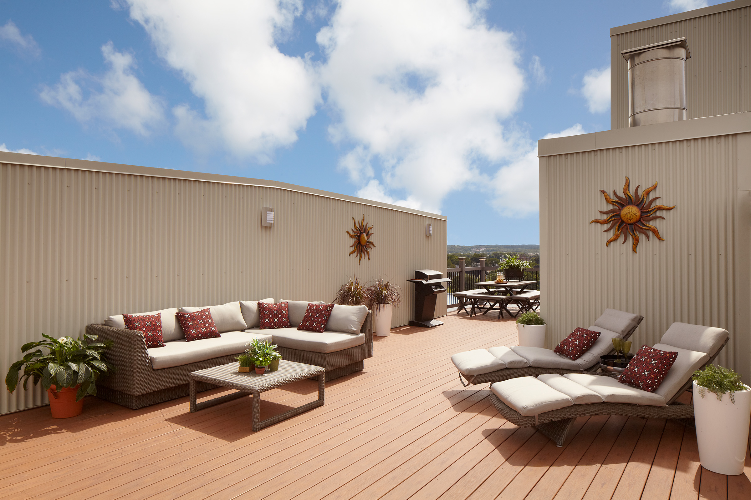 Rooftop deck with wicker sectional seating and lounge chairs accented with red patterned pillows, potted plants, sun-shaped wall art, and a grill, overlooking distant hills.