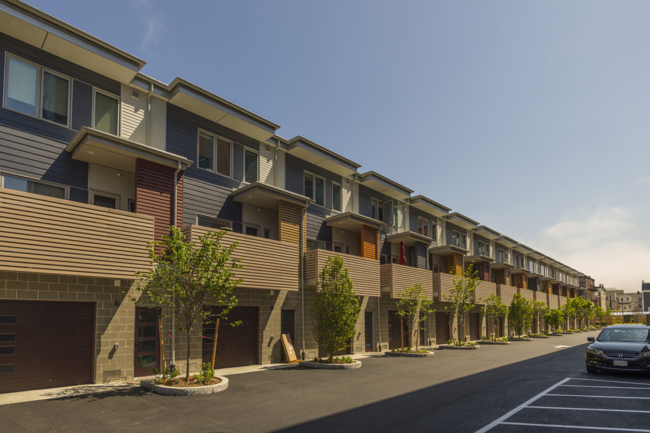 Row of modern townhouses with dark siding, ground-floor garages, wood-slat balconies, and young trees planted along the driveway with a parked car nearby.
