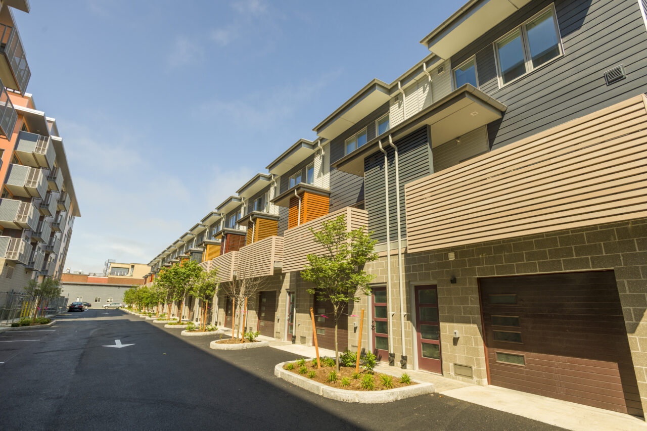 Row of modern townhouses with dark siding and colorful shutter accents, ground-floor garages, and newly planted trees along a paved driveway leading to a parking area.