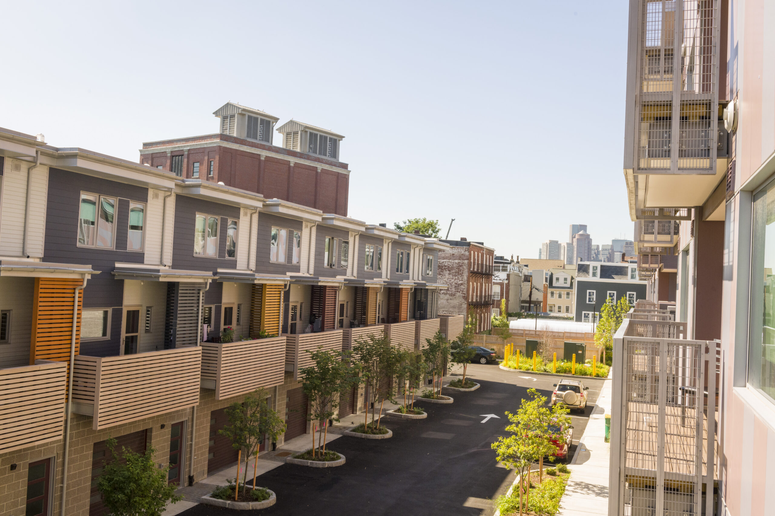 Aerial view of a row of modern townhouses with balconies overlooking a paved driveway lined with young trees, a brick industrial building nearby, and a city skyline in the distance.