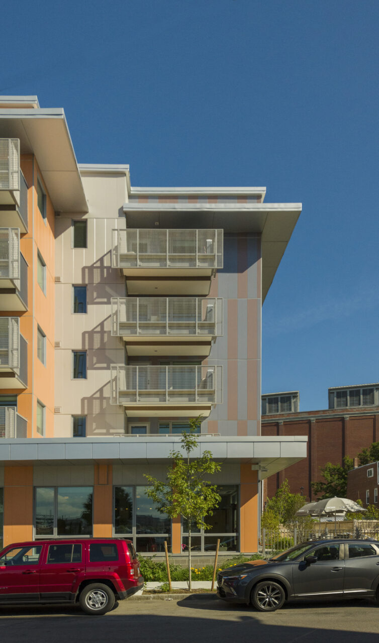 Multi-story apartment building with orange, beige, and gray exterior panels and stacked balconies, above a ground-floor storefront with large windows and parked cars along the street.