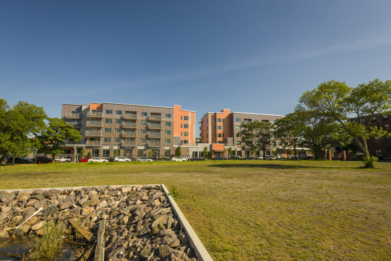 Multi-story orange and gray apartment building seen across a grassy field with a rock-lined drainage channel in the foreground and a parking area near the entrance