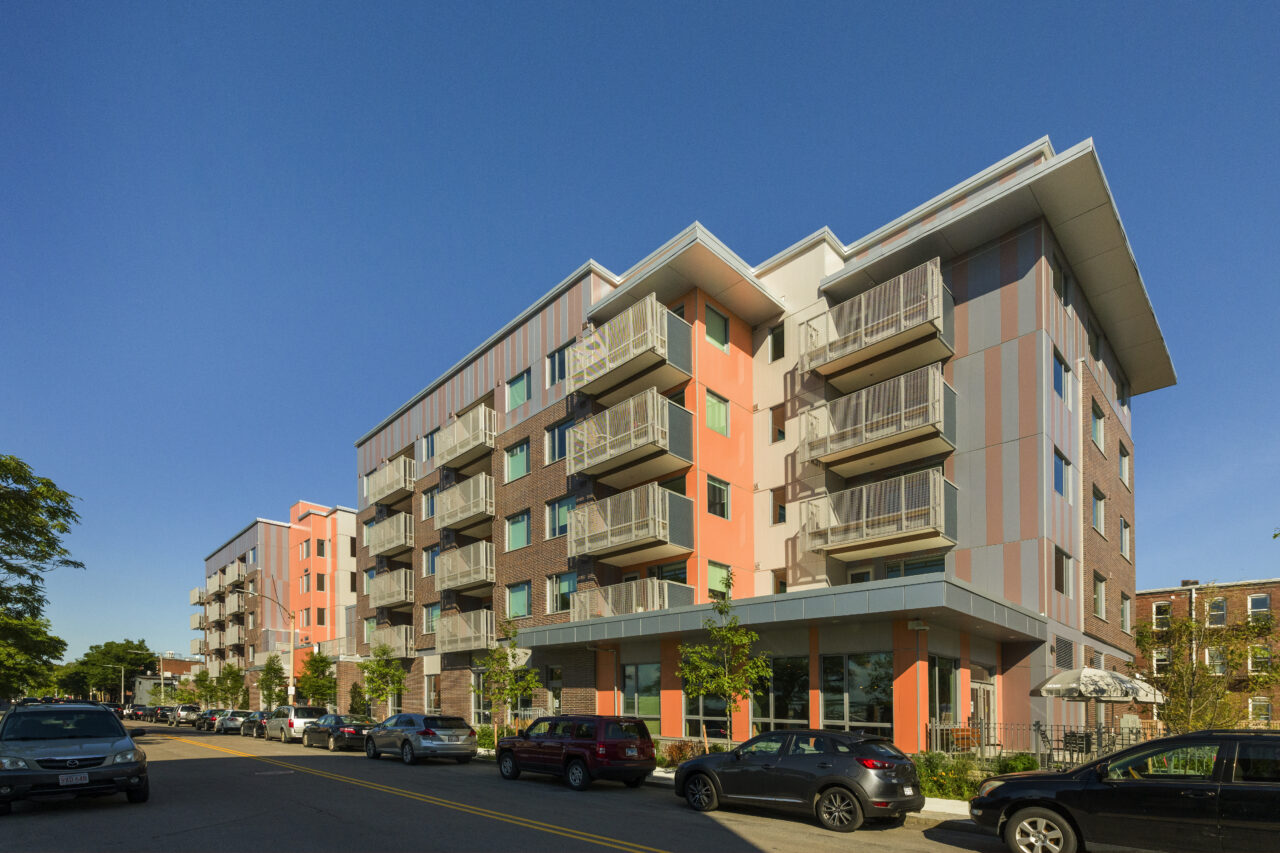 Multi-story orange and gray apartment building with stacked balconies and ground-floor storefronts, along a street lined with parked cars.