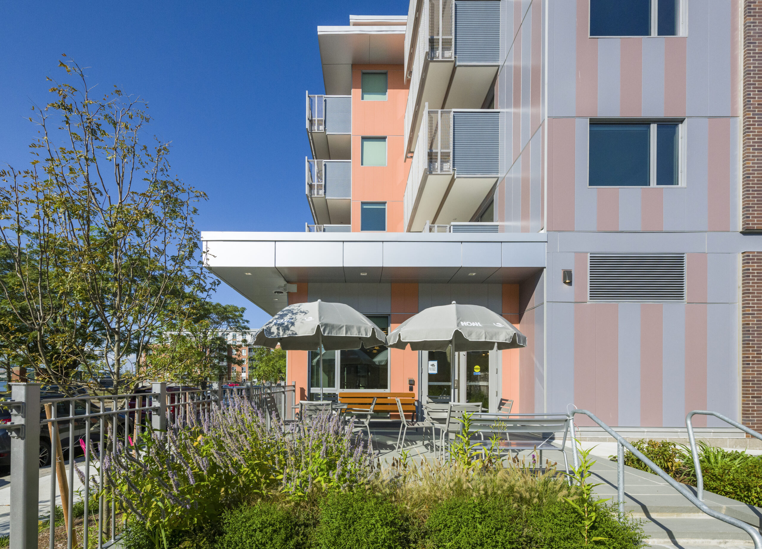 Modern apartment building with orange and gray striped panels and stacked balconies, above a ground-level entrance with an outdoor patio featuring umbrellas, seating, and landscaped plantings.