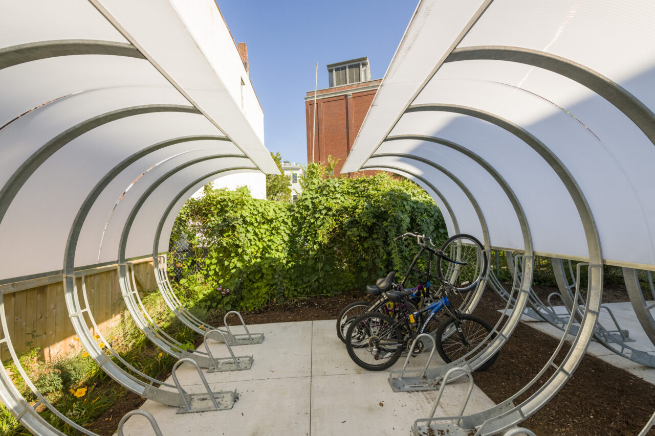 Curved arched bike shelter with translucent panels covering a row of bike racks, with two bicycles parked inside and greenery and a brick building visible beyond.