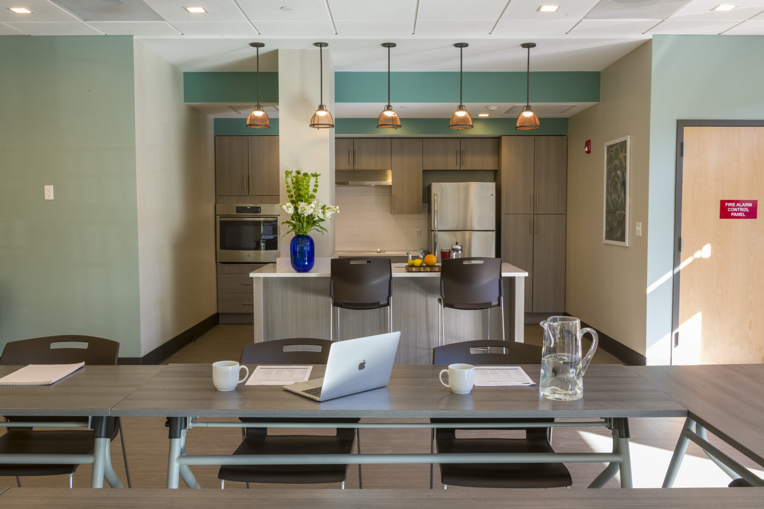 Community room with a long meeting table set with a laptop, mugs, and notepads, leading to a kitchenette with a stainless steel refrigerator, wood cabinets, and pendant lighting over an island.