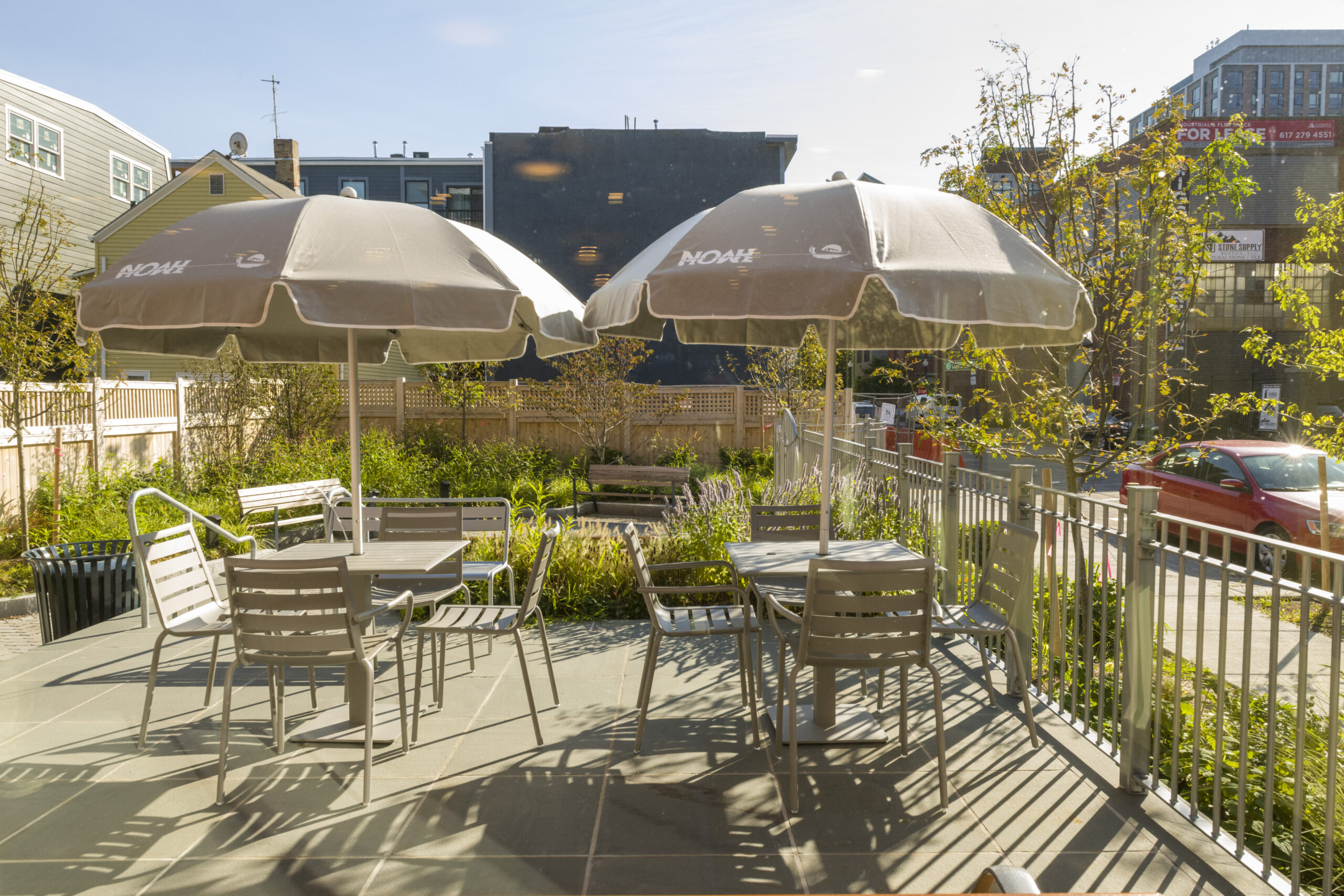 Outdoor patio with metal tables, chairs, and umbrellas on a stone terrace, bordered by a railing and landscaped greenery, with surrounding buildings and a street visible beyond.