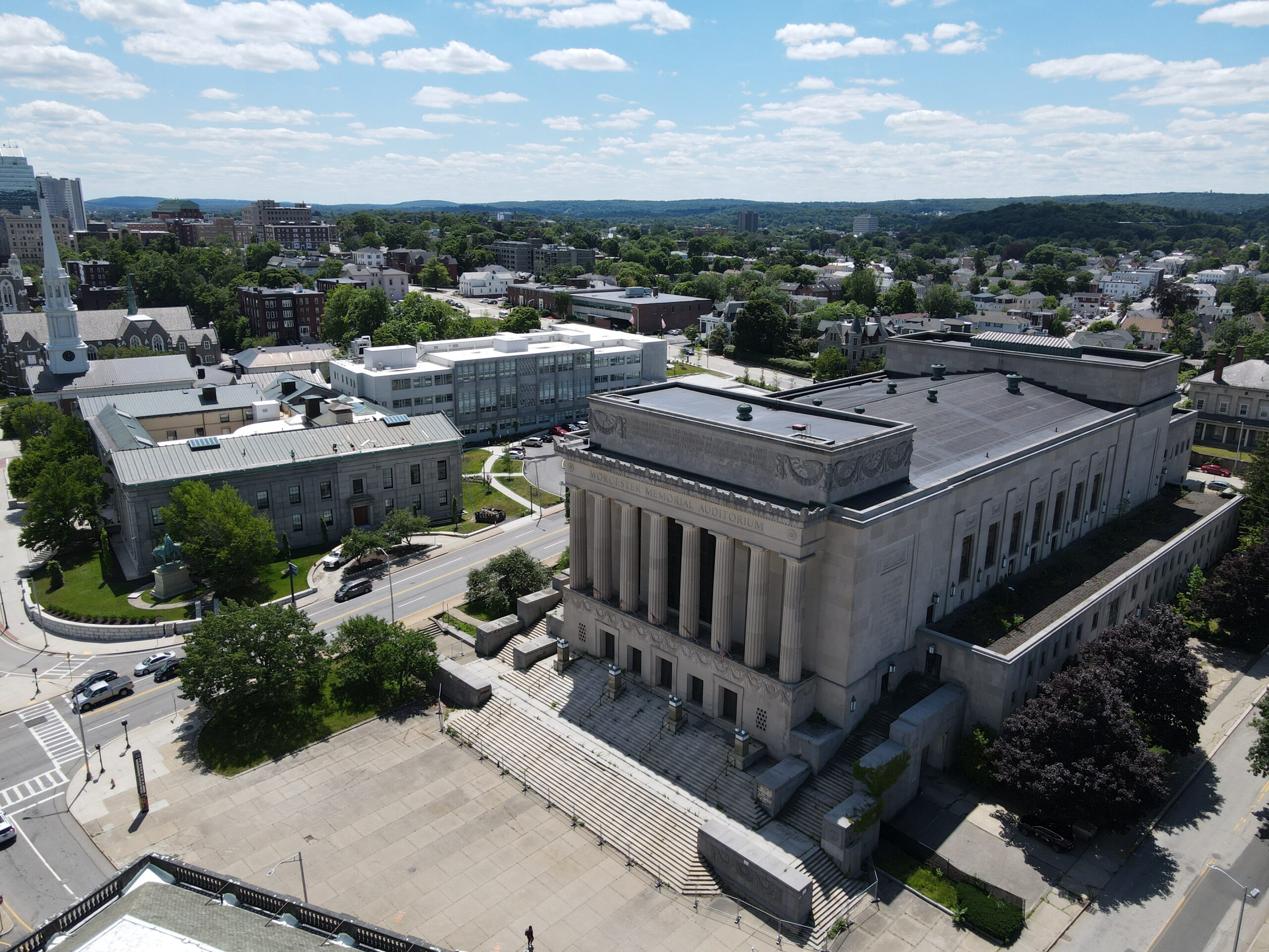 Aerial view of a neoclassical stone building with a colonnaded entrance and wide steps, surrounded by a plaza and city streets.
