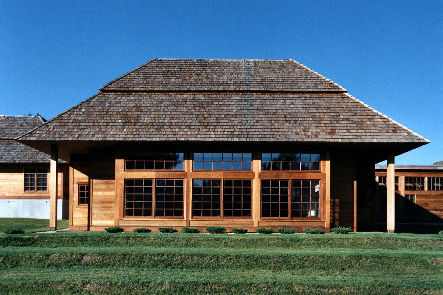 the Yiddish Book Center building exterior, a wood-shingled structure with a deep overhanging roof and large multi-pane windows, set on a grassy lawn.