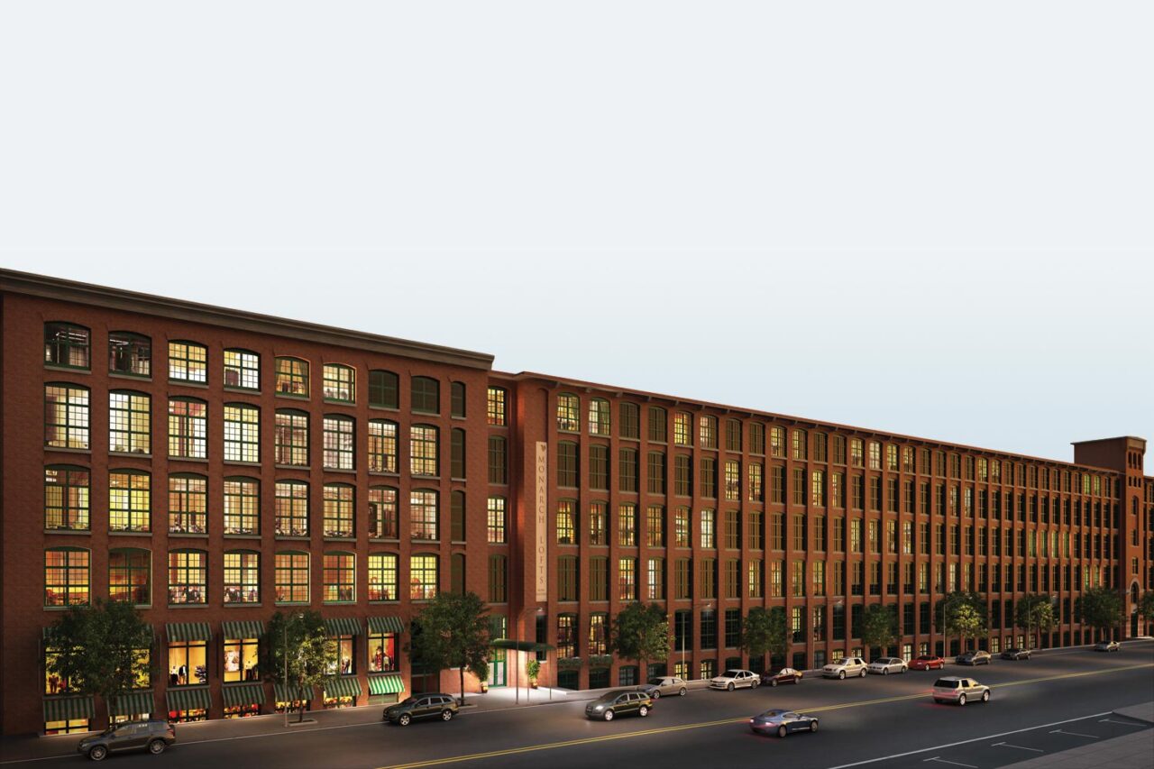 Architectural rendering of Monarch Lofts, a converted brick mill building with rows of arched windows and ground-floor storefronts, stretching along a street lined with parked cars and trees.